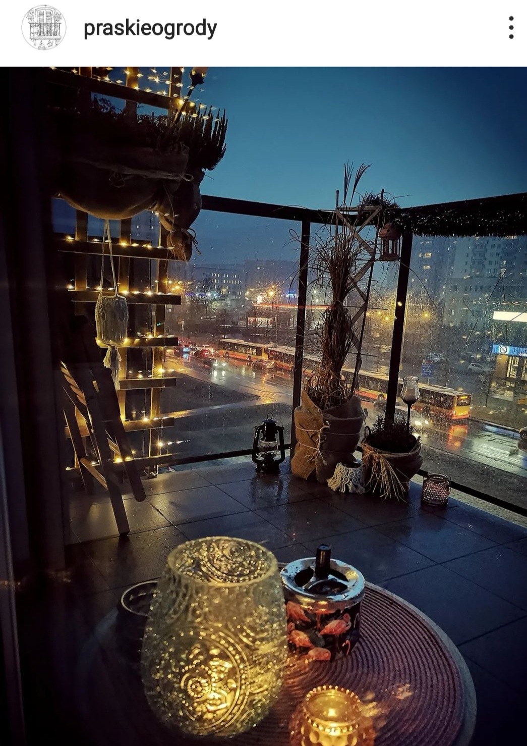 Zadaszony taras na dachu z widokiem na nocne miasto w deszczu, dekoracje roślinne w jutowych workach, lampiony i świecące ozdoby, stolik z popielniczką z motywem flamingów.