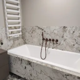 Biała wanna obudowana płytami imitującymi marmur, z miedzianą armaturą i białym grzejnikiem drabinkowym na ścianie w tle.