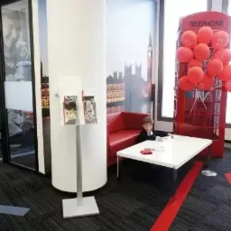 Wnętrze biura z motywem Londynu: czerwona budka telefoniczna z balonami, sofa i stolik, chłopiec w garniturze siedzący przy stoliku, na ścianie fototapeta z Big Benem.