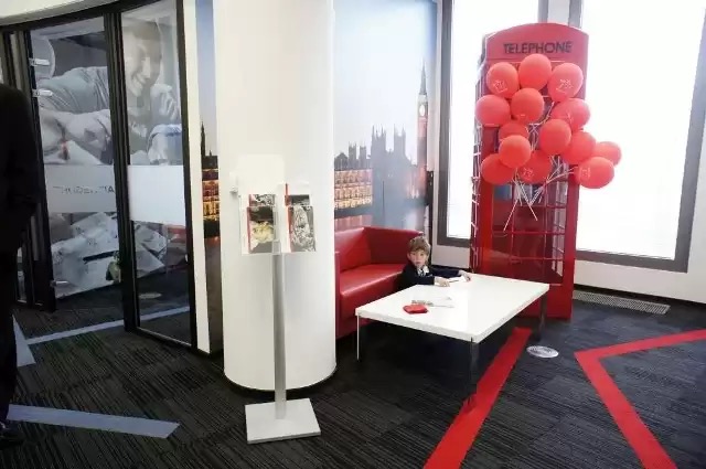 Wnętrze biura z motywem Londynu: czerwona budka telefoniczna z balonami, sofa i stolik, chłopiec w garniturze siedzący przy stoliku, na ścianie fototapeta z Big Benem.