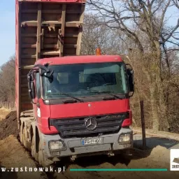 Czerwona ciężarówka Mercedes-Benz z podniesioną skrzynią ładunkową stoi na grząskim terenie budowy; w tle widoczna koparka i drzewa bez liści.