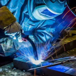 Osoba w masce ochronnej i rękawicach spawalniczych wykonuje spawanie metalu, widoczne iskry i dym w oświetleniu warsztatowym.