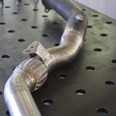 Spawany element rury wydechowej ze stali nierdzewnej z fragmentem elastycznym, widoczne spawy na tle stołu spawalniczego z otworami.