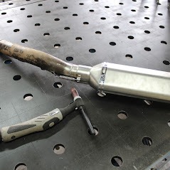 Spoina spawalnicza na rurze wydechowej motocykla, leżącej na perforowanym stole spawalniczym, widoczny uchwyt spawarki TIG.