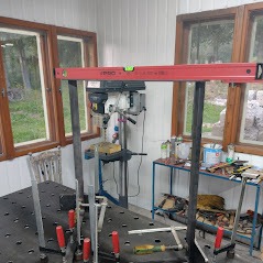 Stalowa konstrukcja spawana na perforowanym stole spawalniczym, poziomowana przy użyciu poziomicy laserowej, w tle wiertarka stołowa i okno z widokiem na zieleń.