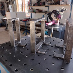 Metalowe konstrukcje spawane, ustawione na perforowanym stole warsztatowym, widoczne narzędzia i elementy wyposażenia warsztatu w tle.