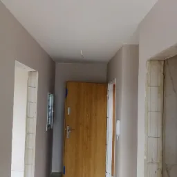 Korytarz w trakcie remontu z świeżo pomalowanymi ścianami w odcieniach szarości, widoczne surowe ościeżnice drzwiowe i częściowo otwarte drzwi wejściowe z okleiną drewnopodobną.