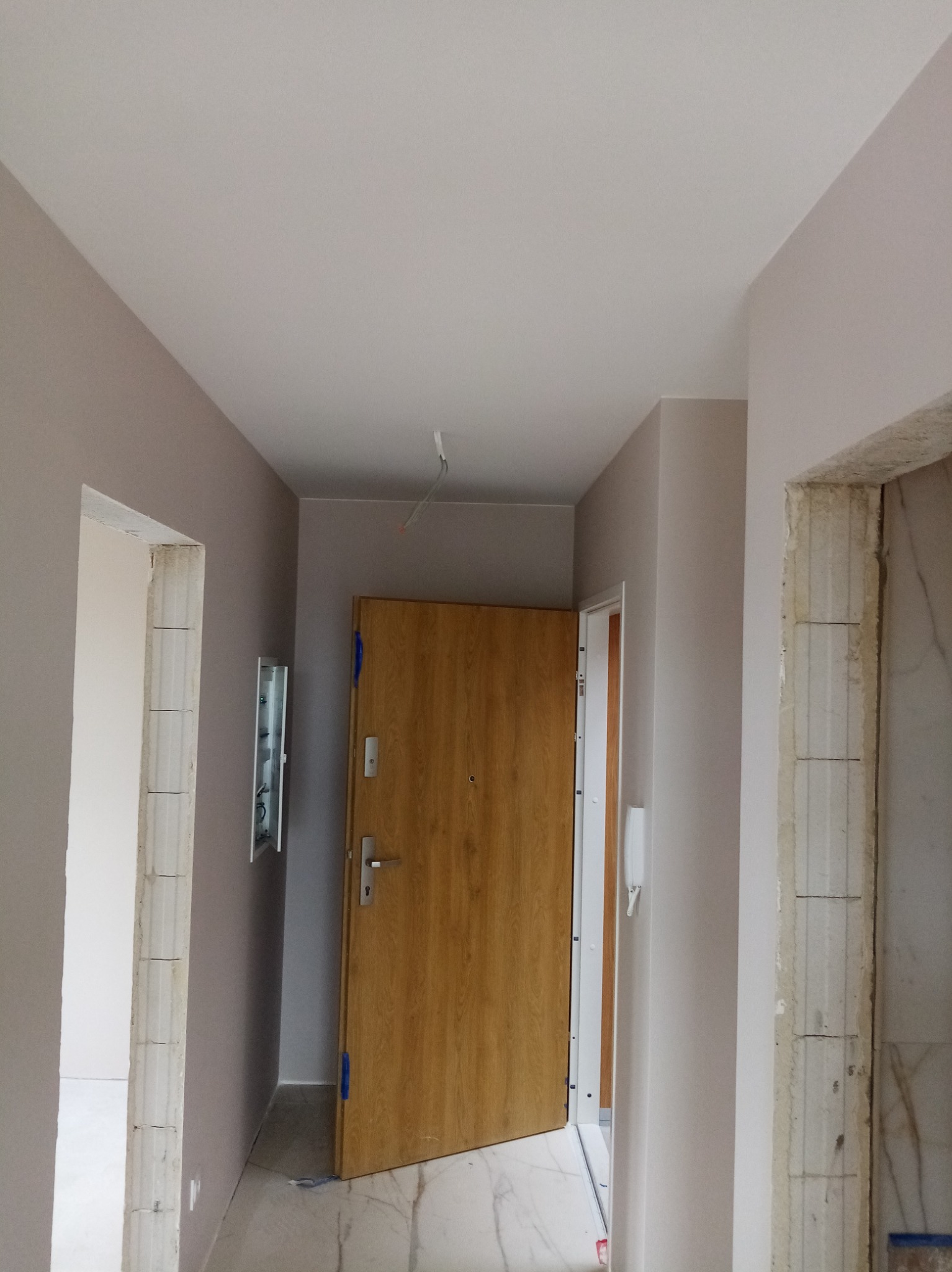 Korytarz w trakcie remontu z świeżo pomalowanymi ścianami w odcieniach szarości, widoczne surowe ościeżnice drzwiowe i częściowo otwarte drzwi wejściowe z okleiną drewnopodobną.