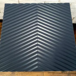 Szczegółowy widok ciemnoszarego, ryflowanego panelu ściennego ułożonego we wzór jodełki, z widocznym fragmentem białej ściany z cegły i drewnianej podłogi.