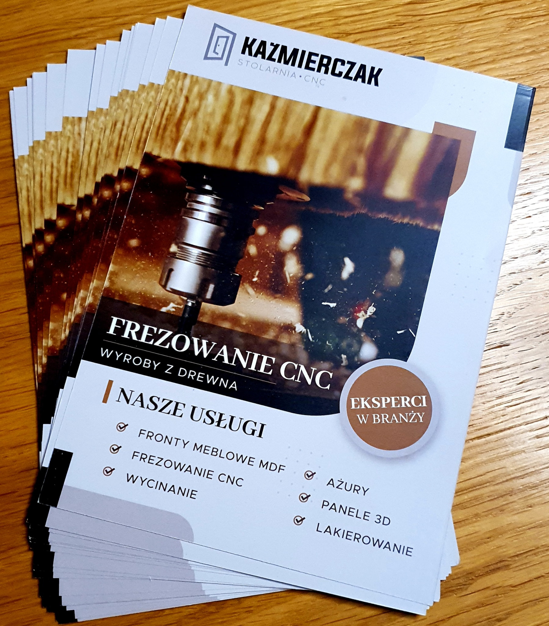 Stos wizytówek stolarni CNC 'Kazmierczak', prezentujących usługi frezowania CNC, fronty meblowe MDF, wycinanie, ażury, panele 3D i lakierowanie.