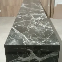 Długi blat z czarnego marmuru z wyraźnym białym żyłkowaniem, na tle ściany z paneli drewnopodobnych i fragmentu grzejnika.