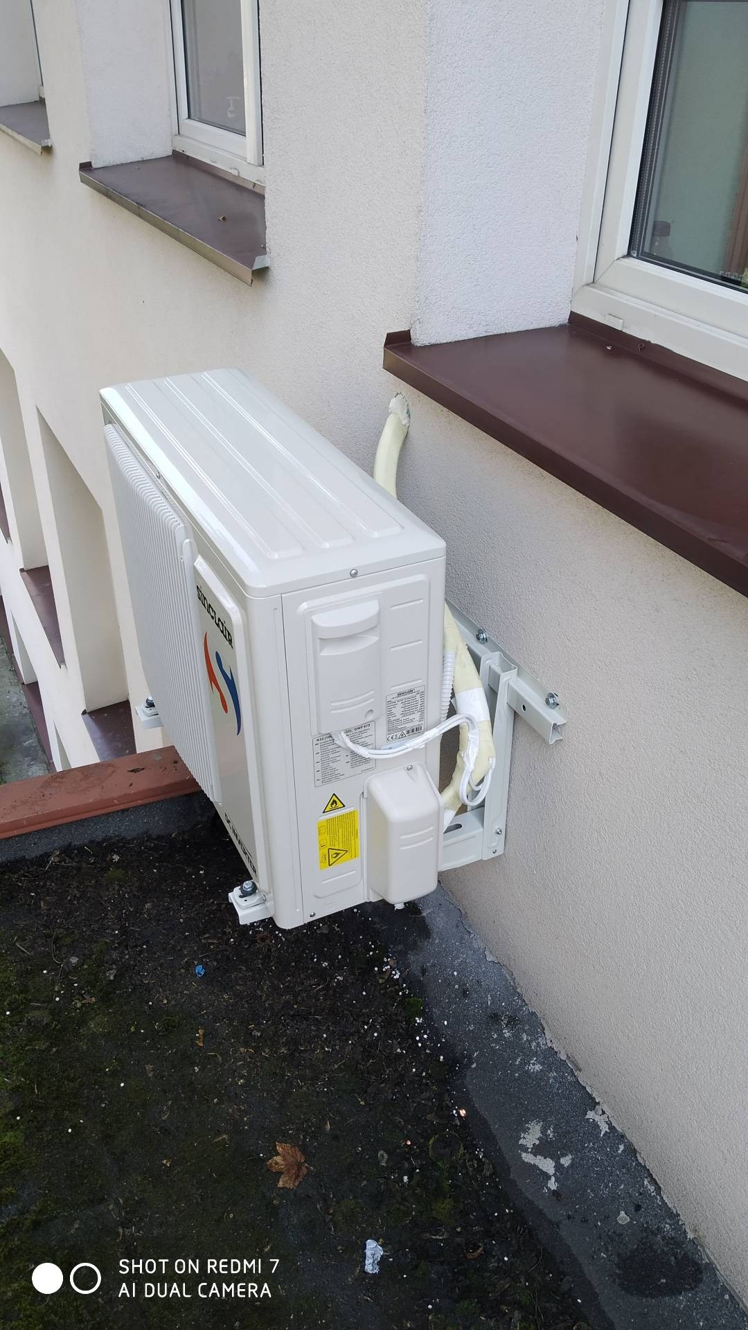 Zewnętrzna jednostka klimatyzacji Sinclair zamontowana na elewacji budynku, widoczne przewody zabezpieczone taśmą, tło stanowią okna z brązowymi parapetami i fragment dachu pokrytego mchem.