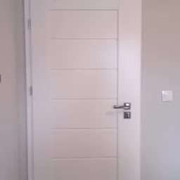 Białe drzwi wewnętrzne z poziomymi frezami, chromowana klamka i zamek, zamontowane w szarej ścianie, widoczny fragment podłogi.