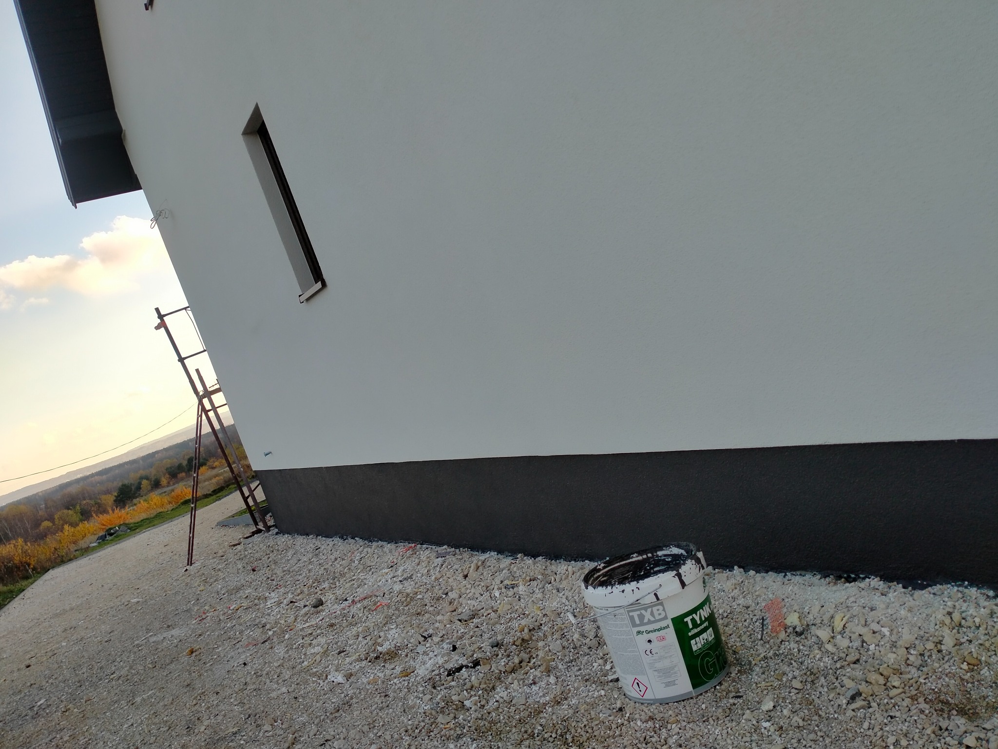 Świeżo pomalowana elewacja domu w kolorze białym z ciemnoszarym cokołem, widoczna drabina oparta o ścianę, wiadro z farbą TXB i kamieniste podłoże.