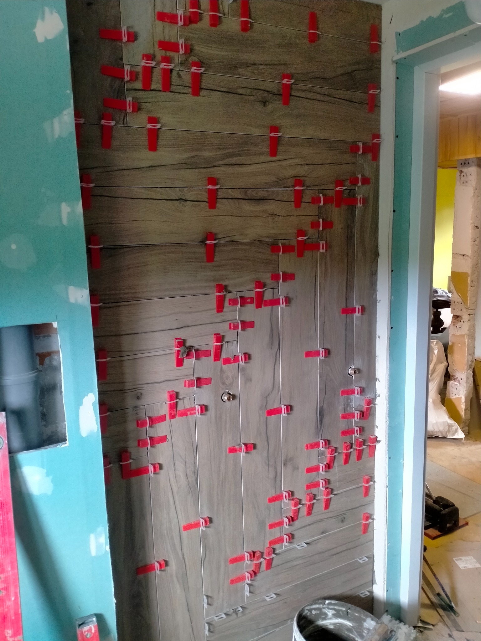 Ściana z płytek imitujących drewno w trakcie montażu, z użyciem czerwonych klipsów poziomujących, widoczne narzędzia i fragment futryny drzwi.