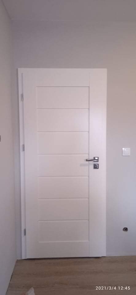 Białe drzwi wewnętrzne z poziomymi frezami, chromowana klamka i zamek, zamontowane w szarej ścianie, widoczny fragment podłogi.