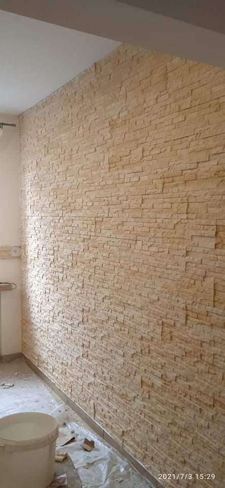 Wnętrze z wykończoną ścianą z dekoracyjnych, beżowych płytek imitujących kamień, widoczny fragment sufitu, podłoga zabezpieczona folią malarską, białe wiadro z farbą w rogu pomieszczenia.