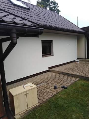 Świeżo pomalowana, biała ściana domu jednorodzinnego z brązową stolarką okienną i rynnami. Na brukowanym podłożu widoczny niski drewniany schowek z narzędziami i wiadro z farbą.