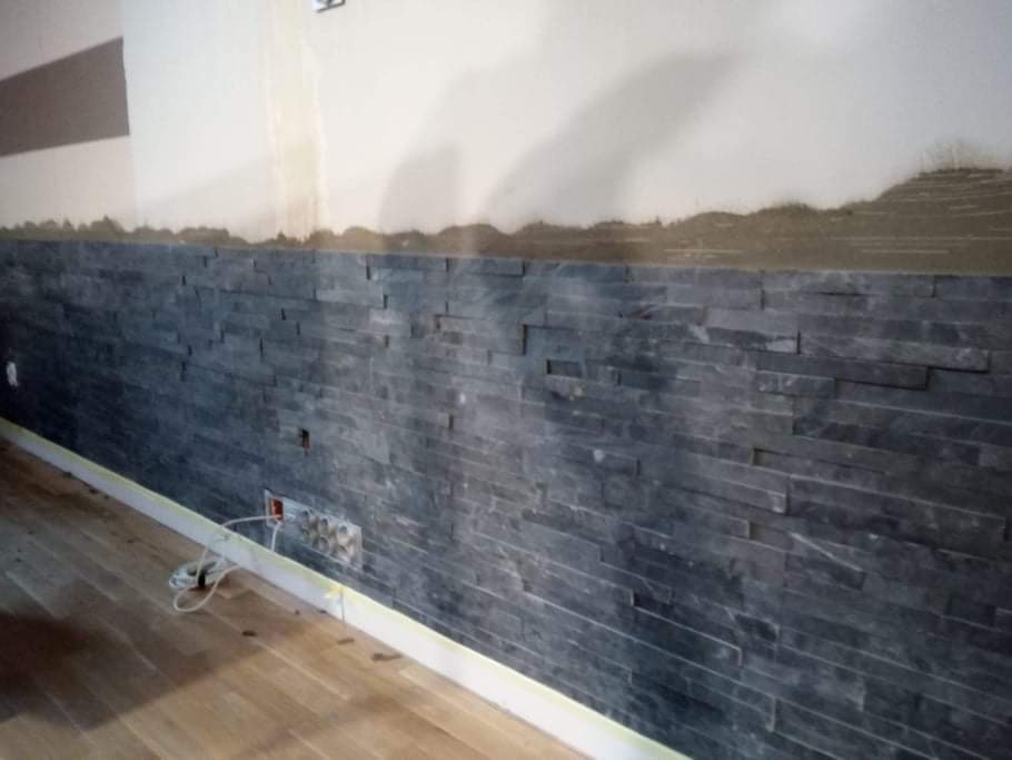 Ściana wewnętrzna wykończona szarą okładziną kamienną, częściowo położona, z widocznymi gniazdkami elektrycznymi i drewnianą podłogą. Górna część ściany surowa, przygotowana do dalszego wykończenia.