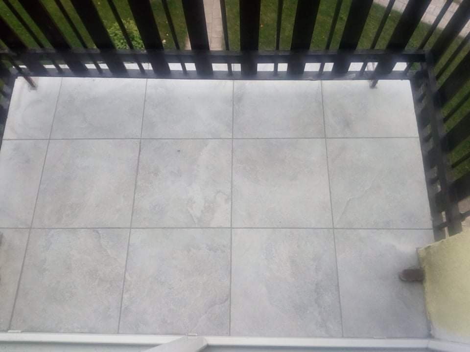 Jasnoszare płytki ułożone na balkonie, widok z góry, ciemne drewniane barierki balkonu, fragment ściany w rogu.