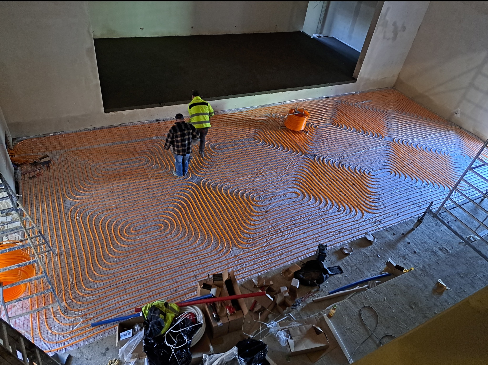 Widok z góry na rozłożoną instalację ogrzewania podłogowego z pomarańczowych rurek na siatce, dwóch monterów w trakcie pracy i częściowo wylaną posadzkę w tle.