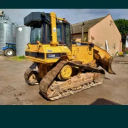 Żółty spychacz marki CAT D5M XL stoi na placu budowy, w tle budynek gospodarczy i silosy zbożowe, widoczne ślady użytkowania na maszynie.