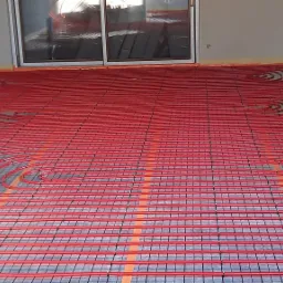 Czerwone rury ogrzewania podłogowego ułożone spiralnie na czarnej siatce montażowej, widoczne okno przesuwne i fragment ściany z taśmą zabezpieczającą wzdłuż krawędzi podłogi.