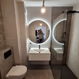 Nowoczesna łazienka z podświetlanym lustrem, kabiną prysznicową typu walk-in i wiszącą toaletą, wykończona płytkami imitującymi marmur w odcieniach szarości i beżu. Widoczne odbicie fotografa...