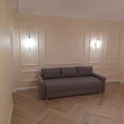 Szara sofa w pokoju z panelami ułożonymi w jodełkę, kremowe ściany z dekoracyjnymi listwami i oświetleniem punktowym, białe drzwi z poziomymi wgłębieniami.