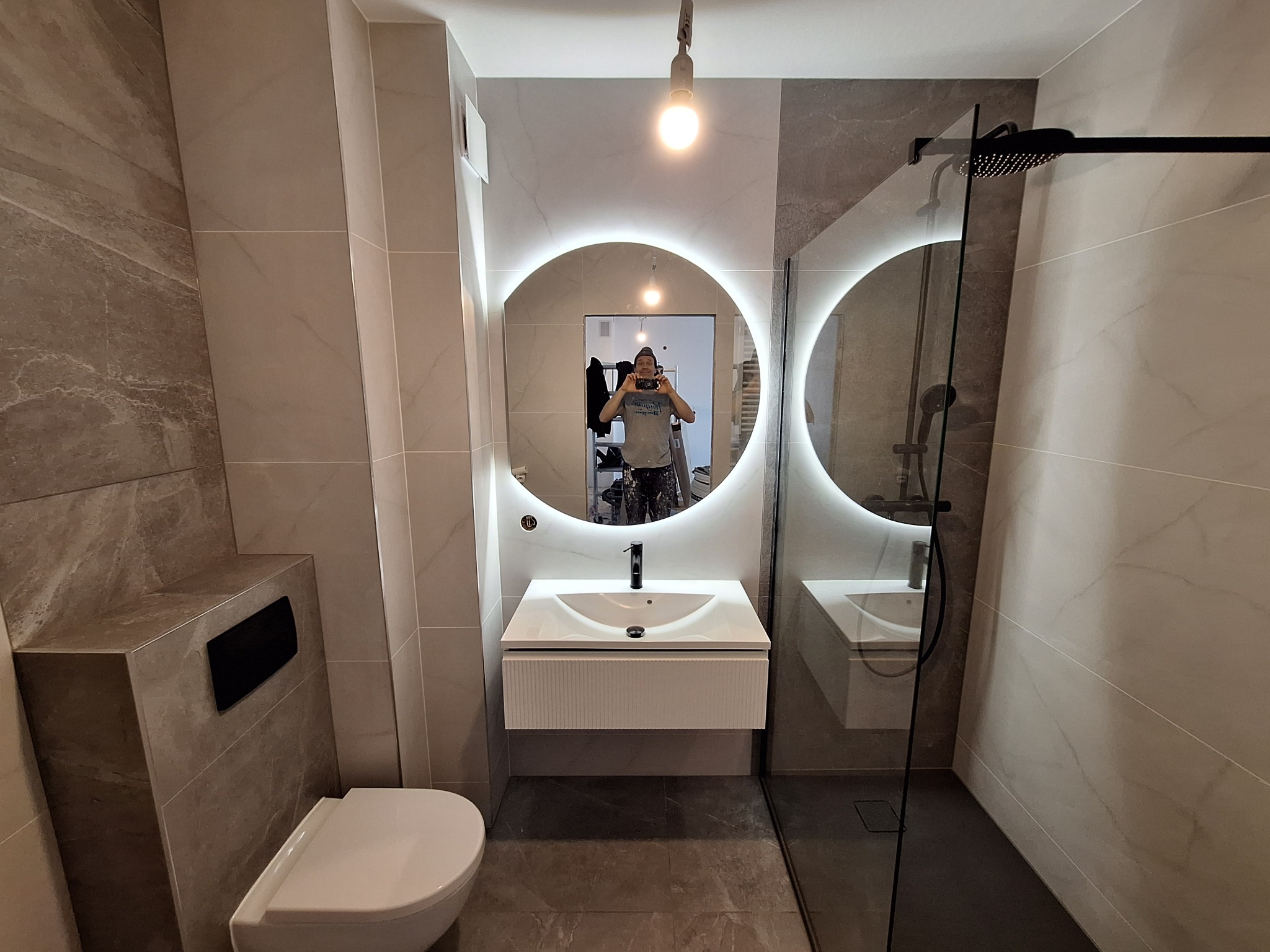 Nowoczesna łazienka z podświetlanym lustrem, kabiną prysznicową typu walk-in i wiszącą toaletą, wykończona płytkami imitującymi marmur w odcieniach szarości i beżu. Widoczne odbicie fotografa...