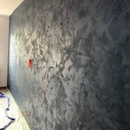 Diman - Ściana z dekoracyjną, szarą fakturą, widoczne wystające kable elektryczne i zabezpieczona folią podłoga podczas prac wykończeniowych.