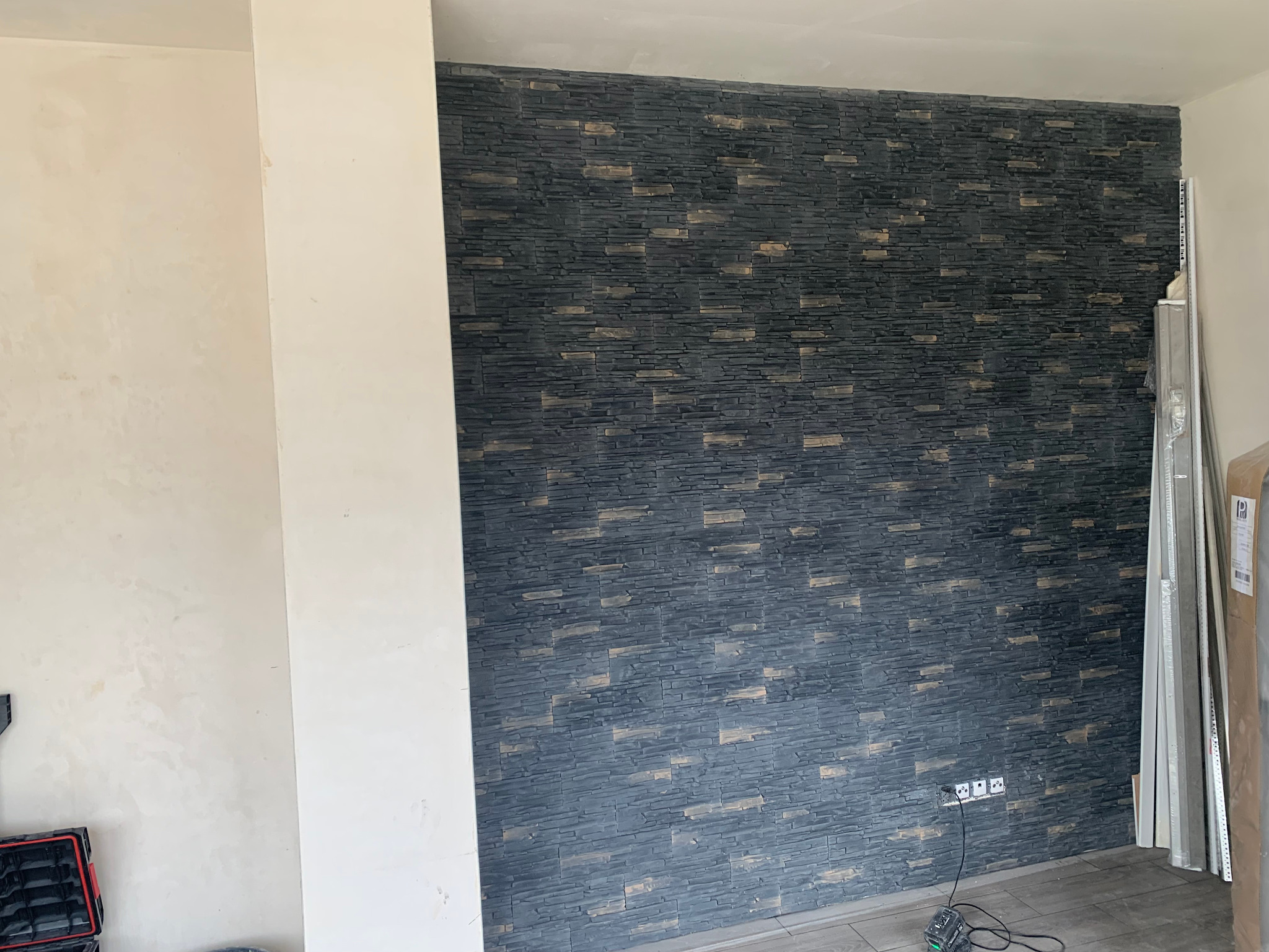 Ściana z dekoracyjnej, ciemnoszarej okładziny kamiennej z beżowymi akcentami, gniazdka elektryczne na dole, fragment ściany przygotowanej do malowania po lewej, listwy przypodłogowe i narzędzia...