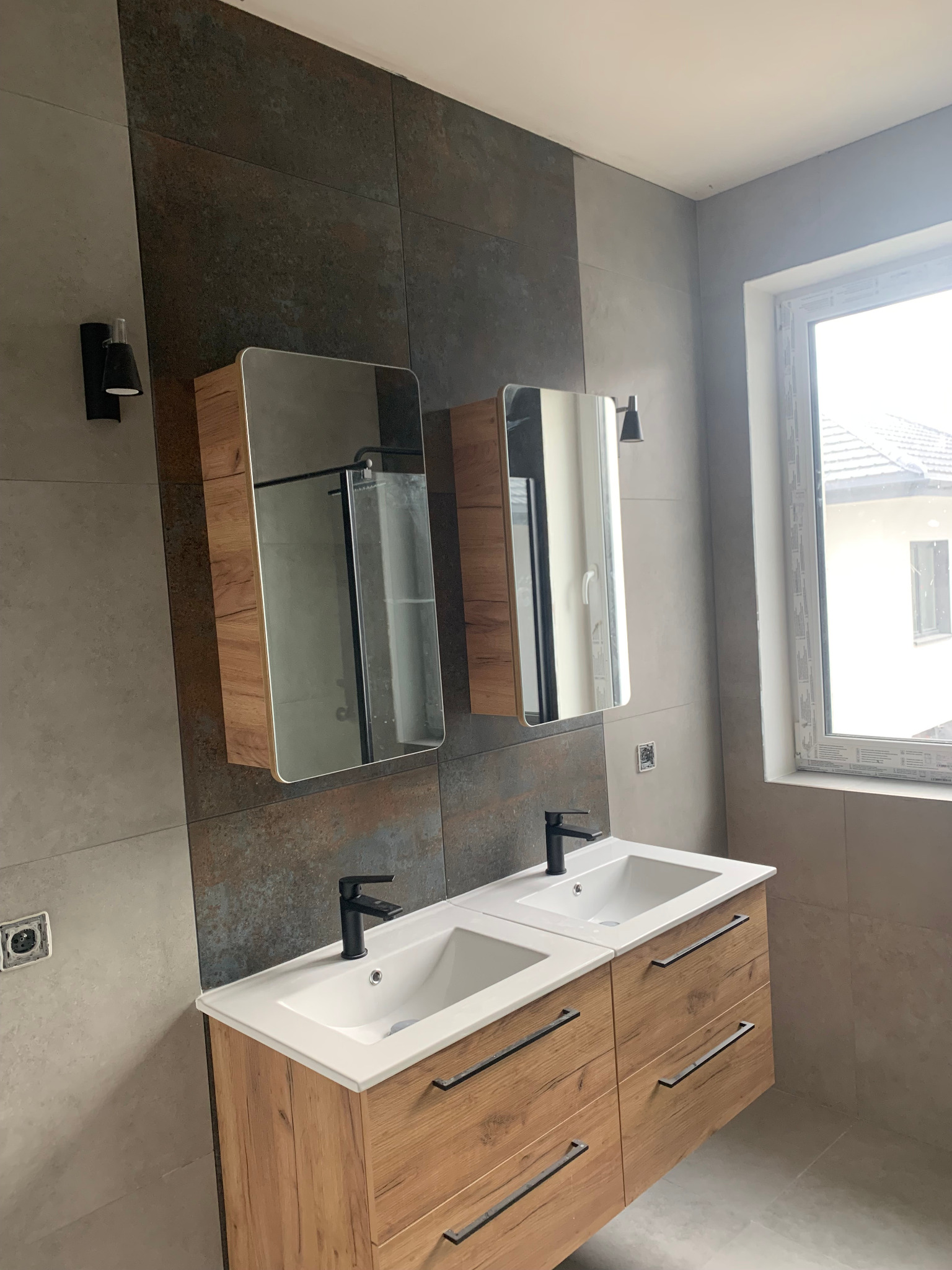 Nowoczesna łazienka z dwoma umywalkami nablatowymi na szafce z drewnopodobnym frontem, czarne baterie, lustra w zabudowie, płytki imitujące kamień na ścianach, czarne kinkiety i okno z widokiem...