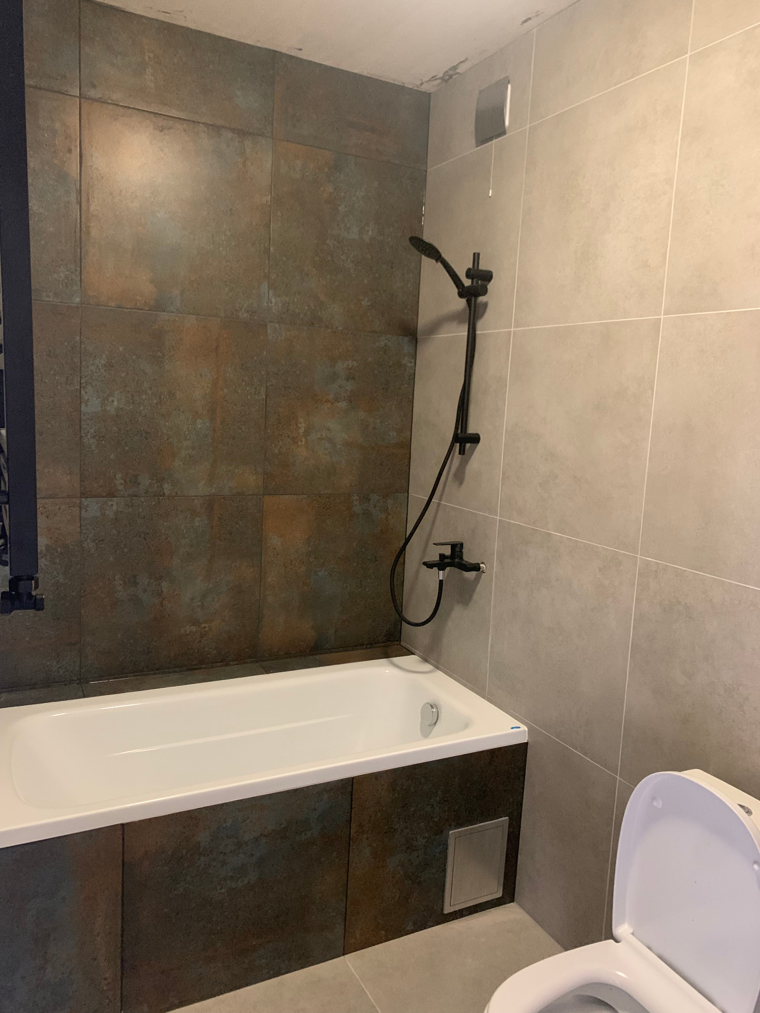 Wykończona łazienka z wanną obudowaną płytkami imitującymi rdzę, czarną armaturą natryskową i jasnymi, gładkimi płytkami na pozostałej części ścian.