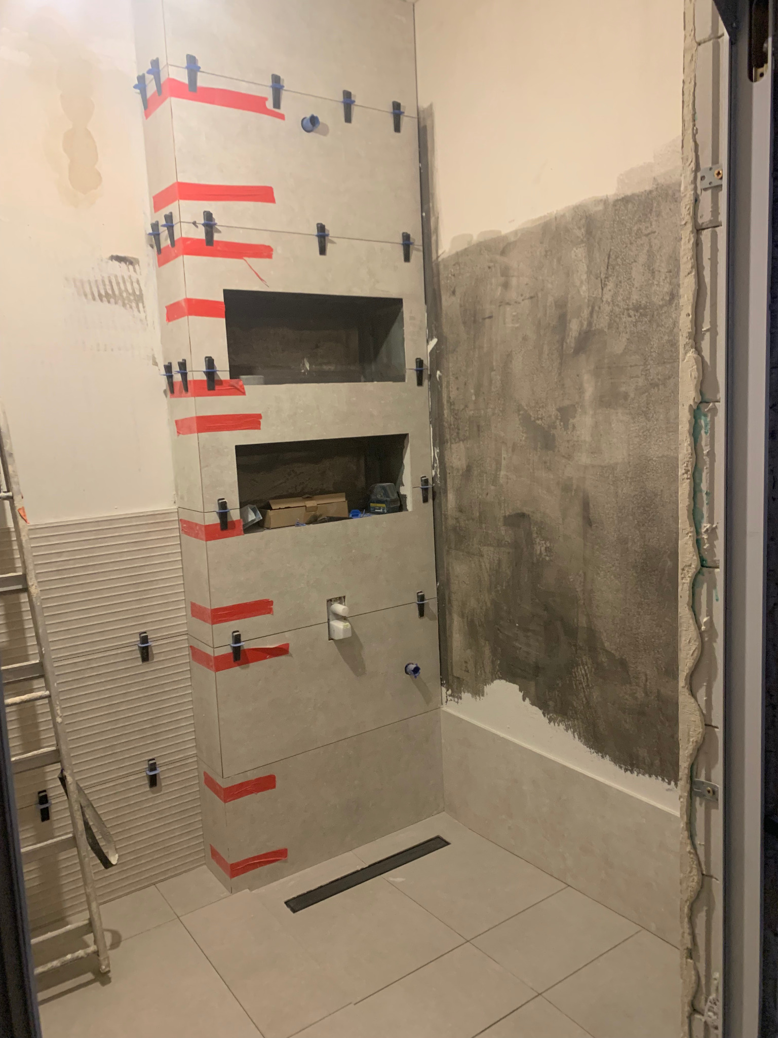 Łazienka w trakcie układania płytek; widoczna wnęka z półkami wyłożona płytkami, poziomowanie płytek za pomocą klipsów i czerwonej taśmy, surowa ściana w tle, drabina oparta o ścianę z ułożonymi...