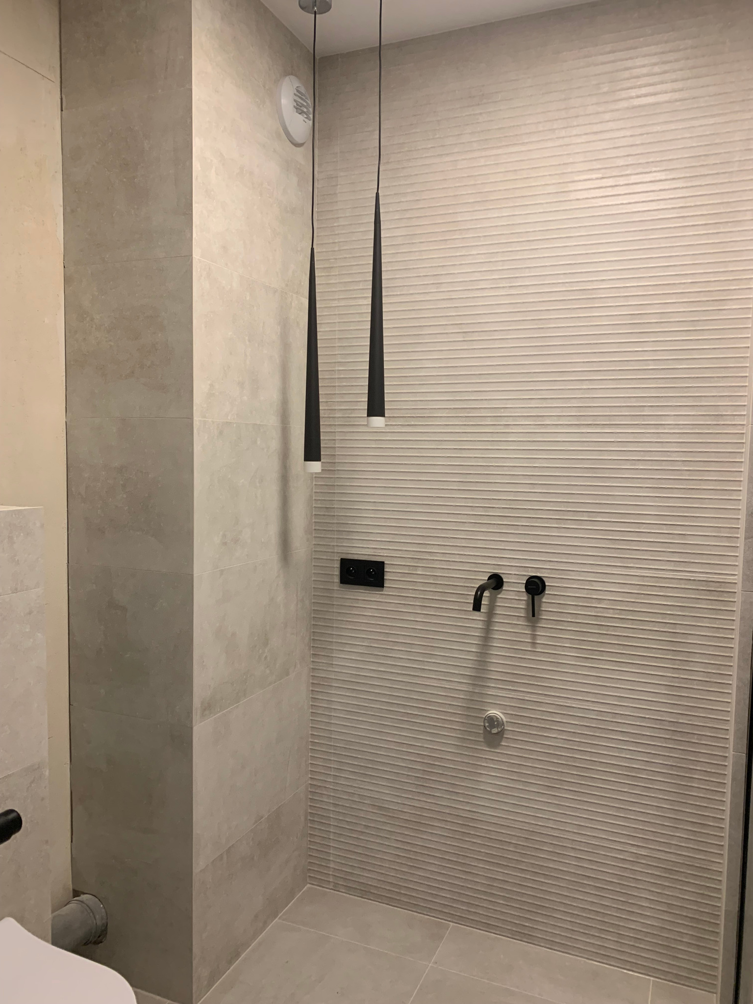 Minimalistyczna łazienka z beżowymi płytkami, czarną armaturą i wiszącymi lampami stożkowymi. Ściana z poziomymi żłobieniami, widoczny fragment toalety po lewej.