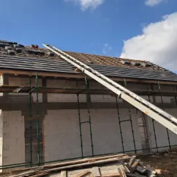 Budowa domu z białych bloczków: częściowo ułożona ciemna dachówka, rusztowania, drabina mechaniczna, błękitne niebo z chmurami w tle.