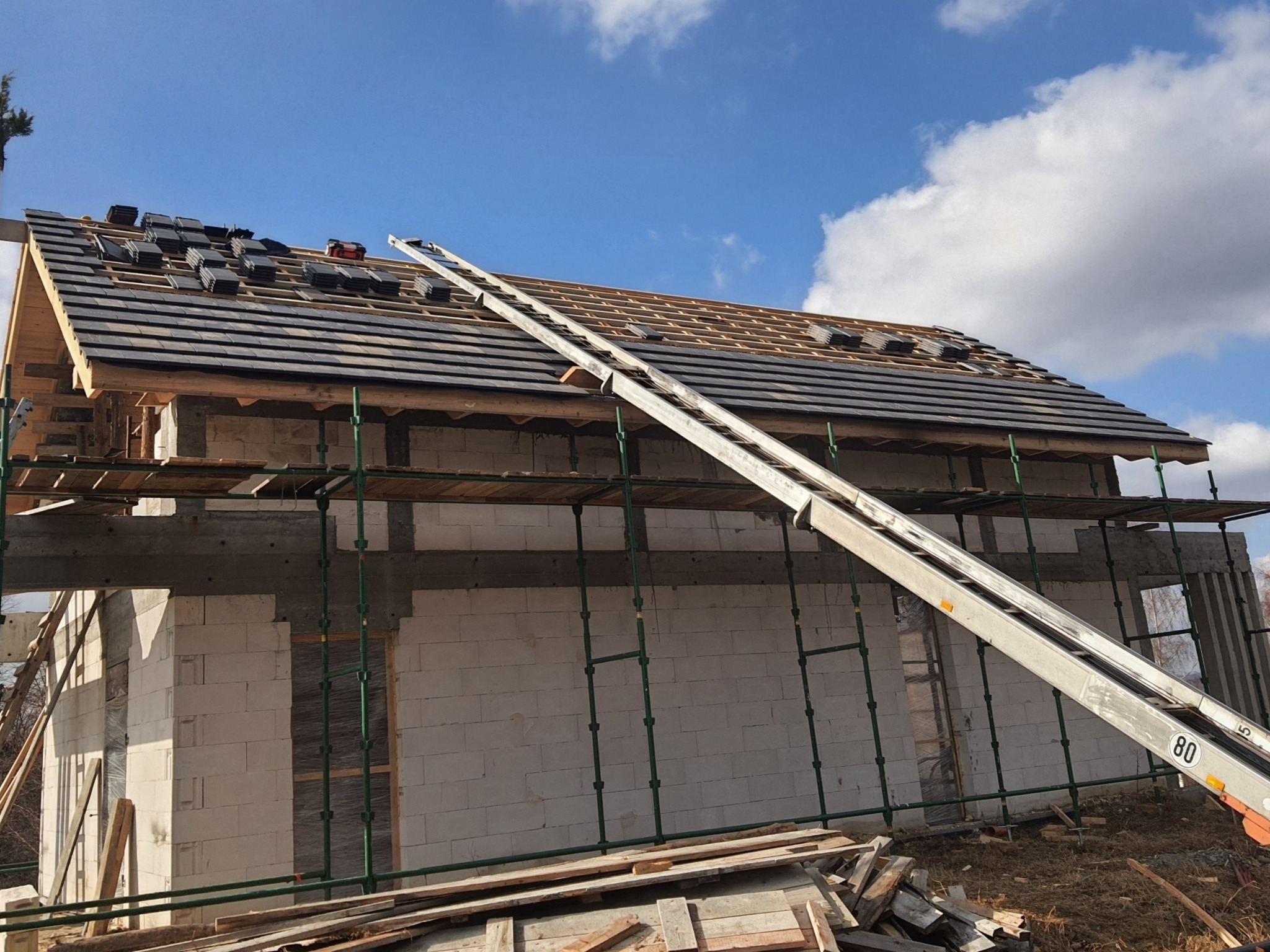 Budowa domu z białych bloczków: częściowo ułożona ciemna dachówka, rusztowania, drabina mechaniczna, błękitne niebo z chmurami w tle.