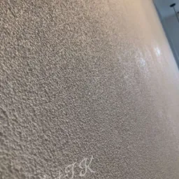 Szara ściana z wyraźną, ziarnistą fakturą, oświetlona z góry, z widocznym odblaskiem światła i delikatnym, odręcznym napisem 'ArtTK' w lewym dolnym rogu.