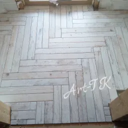 Ułożona podłoga z płytek imitujących drewno w jodełkę, z widocznymi krzyżykami dystansowymi i drewnianymi elementami konstrukcyjnymi u dołu kadru.