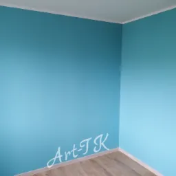 Pomalowane na turkusowo ściany w rogu pokoju z drewnianą podłogą, widoczny biały sufit i listwy przypodłogowe, na ścianie delikatne logo 'Art-TK'.