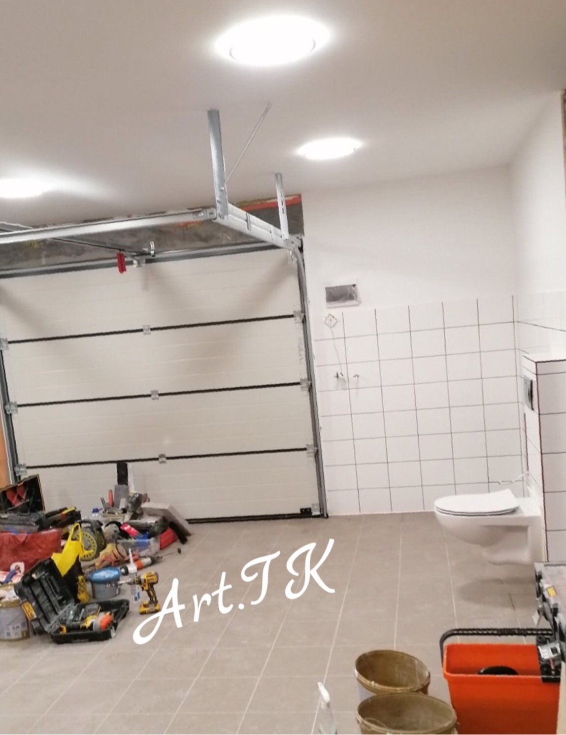 Wnętrze w trakcie prac wykończeniowych z otwartą bramą garażową, widoczne narzędzia, płytki na ścianie i podłodze, oraz toaleta podwieszana.