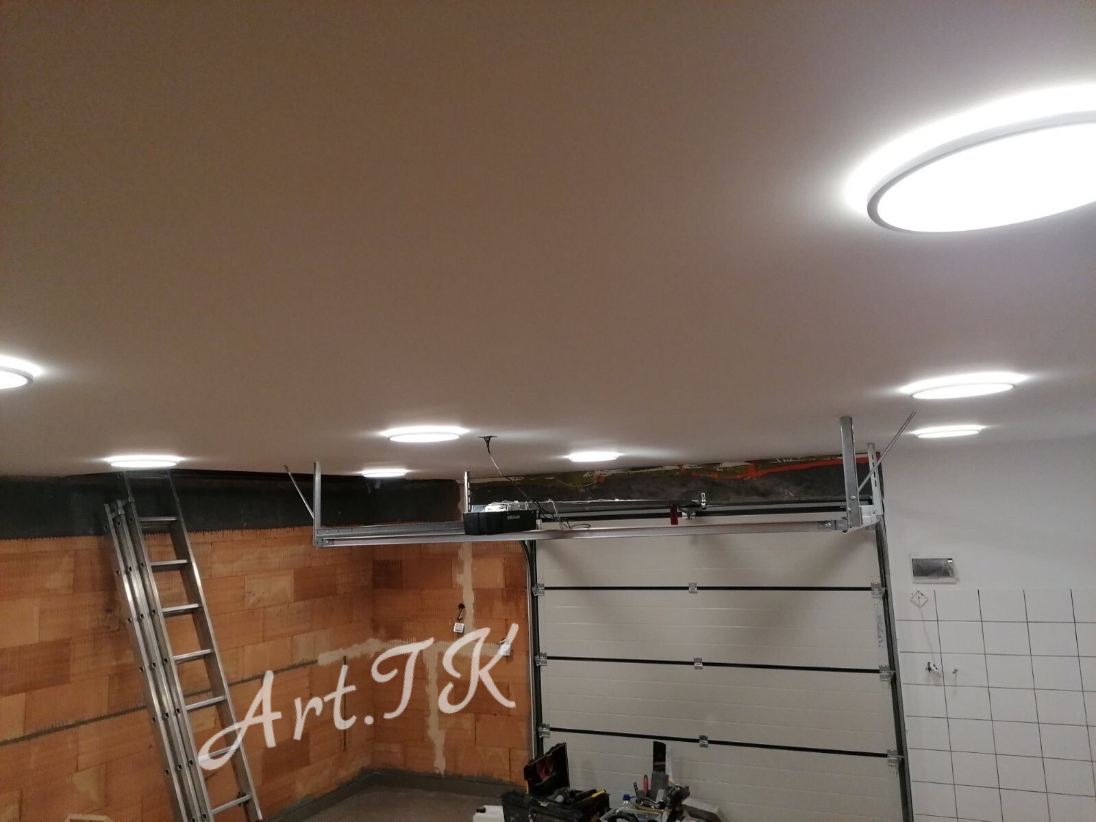 Montaż bramy garażowej segmentowej z napędem elektrycznym, widoczne oświetlenie sufitowe LED, drabina oparta o ścianę z cegły i narzędzia na podłodze.