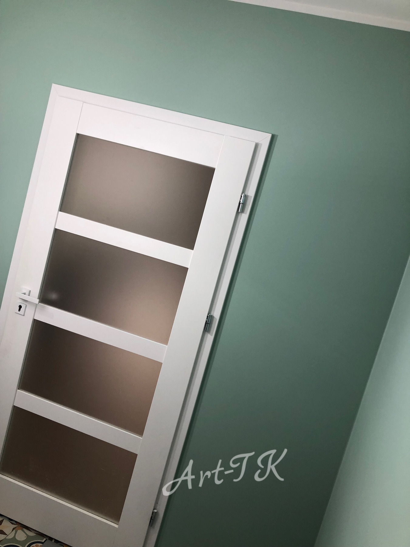 Świeżo pomalowana ściana w odcieniu zieleni z zamontowanymi białymi drzwiami z matowego szkła, widoczne logo 'Art-TK' na ścianie.