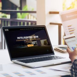taniastronadlafirm.pl - Strony WWW Marketing SEO SEM - Laptop wyświetlający stronę internetową o inteligentnym domu, obok telefon z raportem biznesowym i dokumenty z wykresami, na jasnym stole.