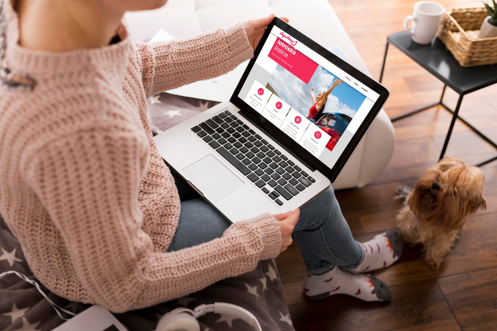 Kobieta w swetrze przegląda na laptopie stronę internetową parkingu lotniskowego, obok siedzi pies rasy Shih Tzu.