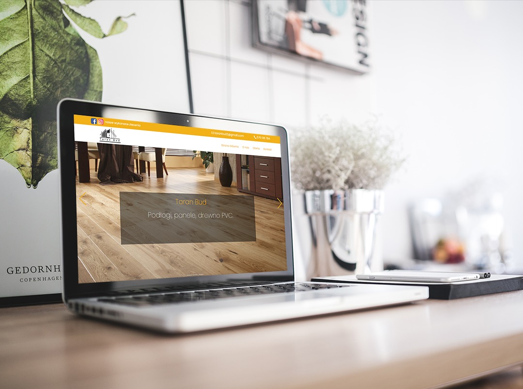 Laptop wyświetla stronę internetową firmy Taran Bud specjalizującej się w podłogach, panelach i drewnie PVC, umieszczony na drewnianym biurku obok smartfona i srebrnego wazonu z kwiatami, w tle...