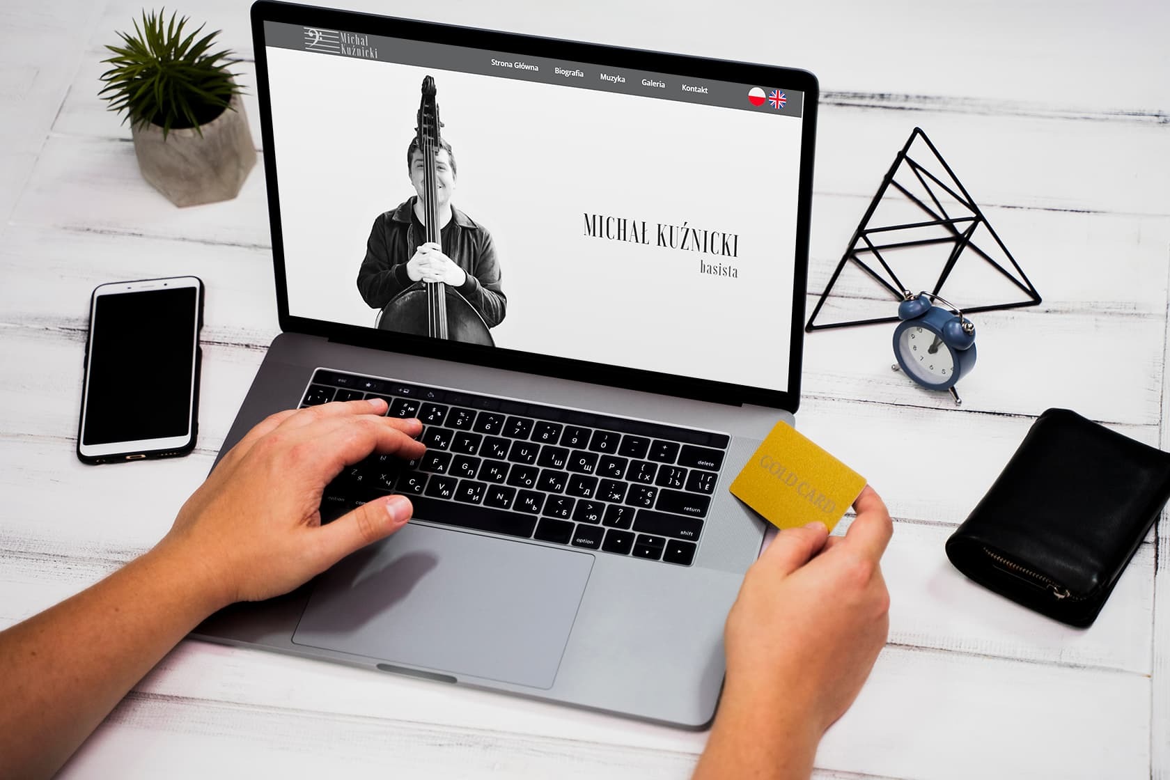 Laptop wyświetla stronę internetową muzyka Michała Kuźnickiego, na drewnianym biurku obok telefon, portfel i złota karta płatnicza w dłoni.