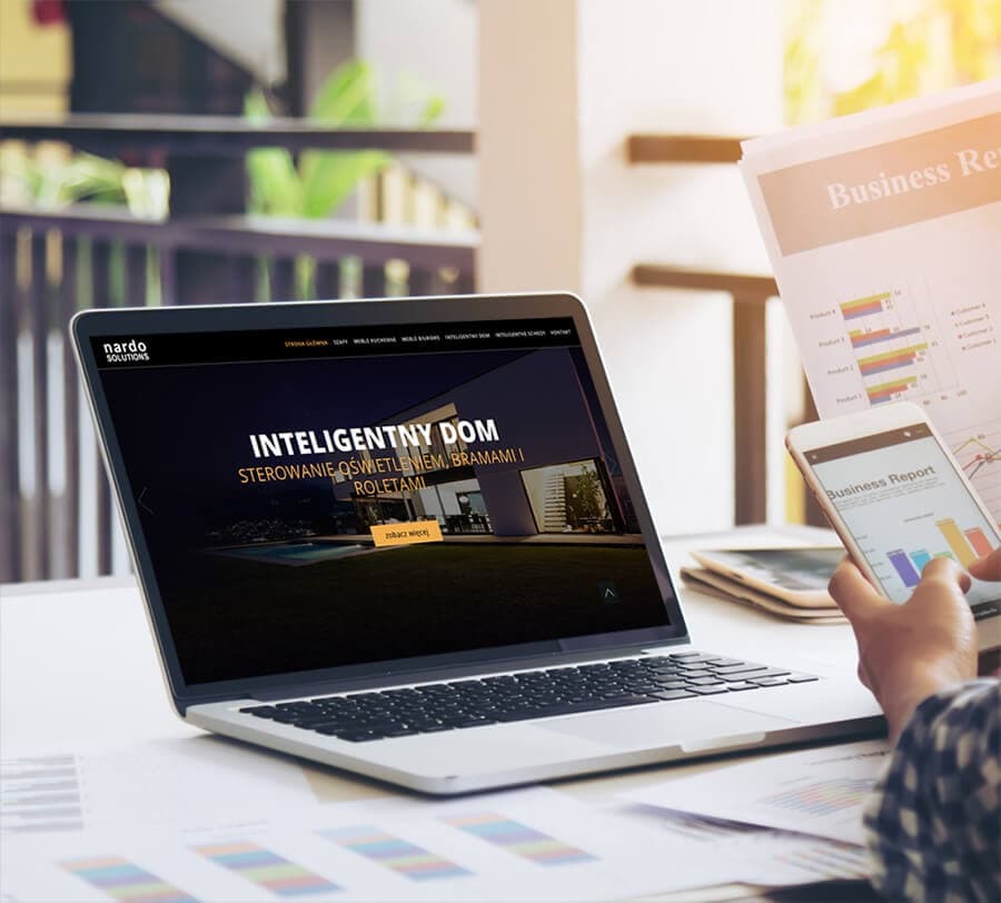Laptop wyświetlający stronę internetową o inteligentnym domu, obok telefon z raportem biznesowym i dokumenty z wykresami, na jasnym stole.