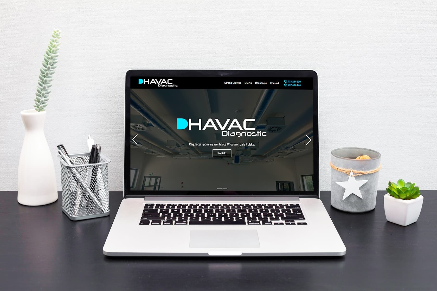 Laptop wyświetlający stronę internetową firmy HVAC Diagnostic, prezentujący wnętrze z systemem wentylacyjnym, obok wazonik z rośliną, organizer na długopisy i świecznik na czarnym biurku.